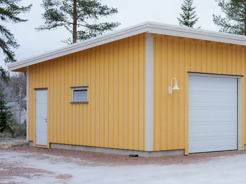 Frittliggende garasje i tre med gul kledning, pulttak, port og dør, på en grusplass ved skogen.